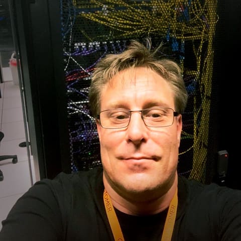Chris Watts, wearing glasses and a black T-shirt, photographed in front of a cabled network rack.