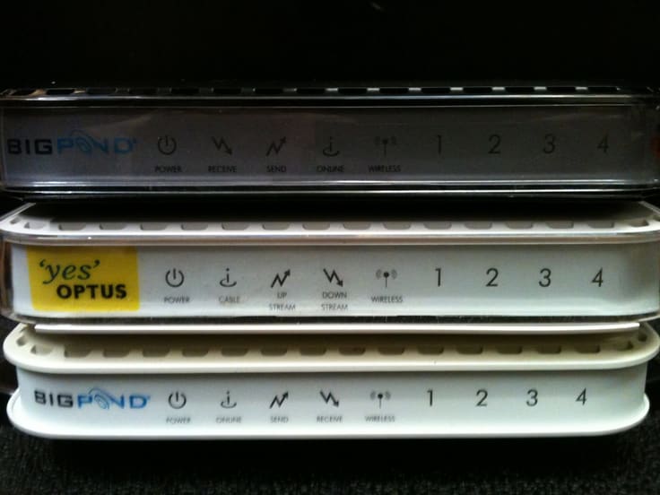 Three stacked cable gateways: a black Telstra BigPond unit on top, a white Yes Optus unit in the middle and a white Telstra BigPond unit below, each with power, cable, send, receive and wireless indicator labels and ports numbered one to four.