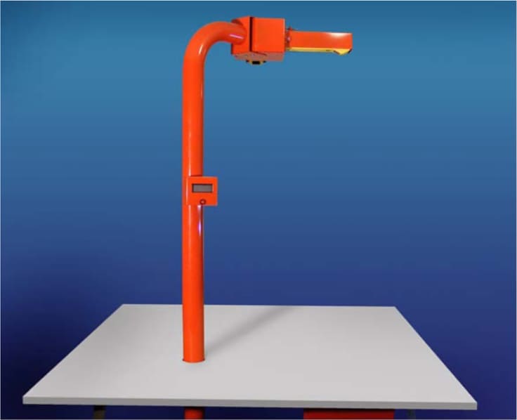 The VCM1250: an orange-red metal arm rising from a white measuring platform, with a small display on the upright and a laser sensor head overhanging the platform.